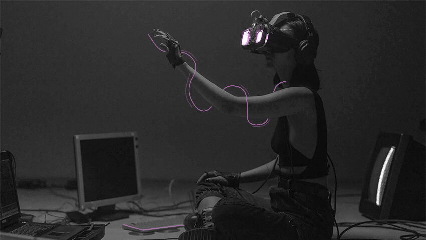 Eine junge Frau sitzt mit VR-Brille vor drei Bildschirmen. Sie ist mit vielen Kabel verbunden und hebt die Hand.